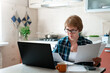 © Marina April - Senior woman working from home kitchen with laptop, documents, finances. Personal budgeting, financial literacy, pension plan, retirement lifestyle. Stress, money anxiety. Ageism in looking for job