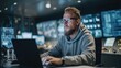© intha - A focused man in glasses and a hoodie works on a laptop in a dimly lit, high-tech control room filled with screens and electronic equipment.