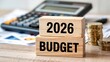 © Design Stock - Wooden blocks stacked with the year 2026 and the word budget in bold black lettering with a calculator and coins in the background