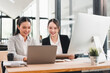 © kenchiro168 - Two Asian businesswomen, smiling and working together at modern office desk, using laptop and digital tablet, with desktop computer and coffee cup, teamwork and collaboration concept