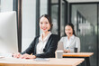 © kenchiro168 - Smiling female customer service representative wearing headset working at modern office desk, with colleague in background, professional and friendly atmosphere
