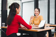 © kenchiro168 - Two Asian women, one in yellow sweater and one in red top, sit at wooden table in modern office, smiling and talking during friendly business meeting