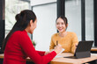 © kenchiro168 - Two Asian women, one in yellow sweater and one in red sweater, sit at wooden table in modern office, smiling and talking, with digital tablet and notebook present