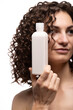 © Alina Zavhorodnii - Portrait of an attractive cheerful long-haired curly-haired girl holding a bottle of soap, shampoo, shower cream, on a white background Mockup