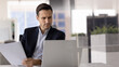 © fizkes - Focused man sits at desk, looks at laptop screen while holding sheet of paper, engaged in task cross-referencing digital and printed information, looks concentrated, busy in data analysis, or planning