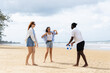 © Sumala - Three people enjoy summer outdoor activities on beach with people playing ball and relaxing under cloudy sky near ocean shore