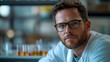 © TANZILA - A handsome male scientist wearing glasses and a beard sits in a modern laboratory, confidently looking directly at the camera, surrounded by scientific equipment and laboratory machinery.