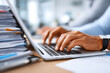 © jantavong - Busy Office Worker Typing on Laptop Surrounded by Stacks of Paperwork in a Modern Workspace