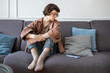 © ingoodtime - Thoughtful woman sadly looking on phone sits on couch waiting call or message feeling loneliness, lost. Melancholic girl alone on sofa suffers uncertain, emotionally distant mobile phone disconnected.