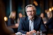 © Tenalxp - A mature man with glasses and a beard sits confidently at a table in a professional setting, with blurred people and warm lights in the background.
