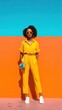 © hack - A confident woman in a yellow jumpsuit and white sneakers stands against a vibrant orange and blue wall, accessorized with round sunglasses and a small blue purse.