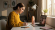 © ikhsan - Focused female student engaged in e-learning, using a laptop for a virtual classroom webinar and writing notes at her sunlit desk at home.
