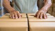 © nunoi - An elderly person's hands rest on several cardboard boxes, conveying themes of labor, care, and the passage of time in a warehouse environment.