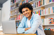 © Home-stock - Portrait of happy African American male student sitting in campus library with laptop, preparing to exam, searching information, looking and smiling at camera