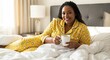 © Milza Mariano - Woman in yellow pajamas with cartoon print holding white mug in bed with white sheets smiling