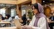 © Y - A young woman in a hijab works diligently on her laptop in a modern, open-plan workspace.