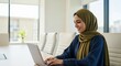 © Fajar - A focused Muslim woman in a hijab works diligently on her laptop, immersed in her professional tasks within a modern setting.
