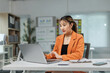 © Crystal - Young Asian businesswoman concentrating on her work, typing on a laptop at her desk in a bright, modern office environment, surrounded by documents and office supplies