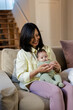 © Wavebreak Media - Mother holding baby girl wearing green onesie while sitting on beige couch in living room