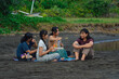 © agratitudesign - Sitting beside a docked boat and a still pond, four girls enjoy their food and drinks on a mat, surrounded by sand, trees, and plants in a calm morning setting.