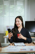 © Kieng - A smiling young woman reviews documents at her desk, with a laptop, calculator, and graphs on a monitor in the background, signifying business analysis.