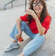 © olezzo - Portrait of a beautiful latino brunette in a red t-shirt and glasses sitting on the steps in a summer city