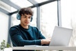 © Prostock-studio - Concentrated young european man wearing headphones is intently using laptop while seated at modern workspace with large window in the background