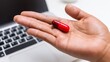 © Marin Zhu - Hand holding a red capsule above a laptop keyboard, symbolizing health and wellness, with a modern workspace in the background, showcasing a blend of technology and medicine