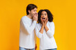 © Prostock-studio - Gossips concept. Boyfriend whispering secret to his surprised afro girlfriend while they standing together yellow background with empty space