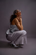 © Seventyfour - Vertical studio shot of young woman crouching in fashion photoshoot, wearing metallic outfit and decorative headpiece, tilting head upward with hands near face posing against plain background