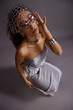 © Seventyfour - Vertical portrait of young multiethnic woman wearing metallic accessories and silver dress, looking into camera while adjusting eyeglasses, curly hair styled with decorative beads, fashion photoshoot