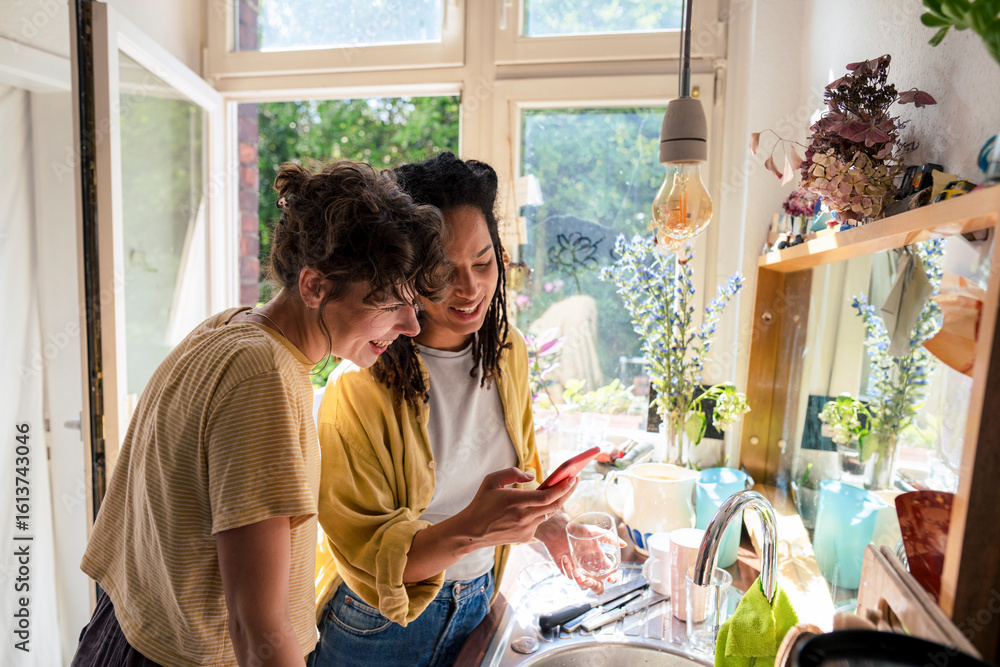 Smiling woman standing near friend using smart phone in kitchen at home