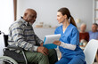 © Prostock-studio - Pretty young woman in workwear nurse helping black senior man in weheelchair filling papers, cheerful elderly african american male patient signing documents at nursing home