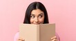 © SAYERA - A young woman smiles while reading a book against a pink background