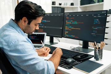 Smart IT developer writing data memo's software development on paper's coding on laptop and two pc screen, creating application program update version online website system at modern office. Surmise.