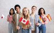 © Prostock-studio - International exchange program and study together. Smiling young multi ethnic students looking at camera and holding flags of their countries, on white brick wall background, copy space, studio shot