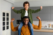 © Prostock-studio - Black preteen boy on his dad's shoulders triumphantly raises his fists in the air, as they both flex their biceps, displaying strength and bonding at home