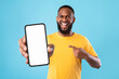 © Prostock-studio - Handsome happy black guy pointing at smartphone with blank screen on blue studio background, mockup for website or new mobile app. Cellphone display template. Space for advertisement