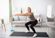 © Prostock-studio - Home workout concept. Beautiful mature lady in sportswear doing squats on yoga mat indoors, free space. Positive senior woman exercising her legs on domestic training during coronavirus isolation