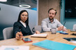 © BullRun - Professional woman reaching for phone while writing during business meeting, surrounded by digital devices, notebooks and colleagues, embodying multitasking and connected workflow