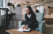 © BullRun - Young woman in glasses looking at camera with soft confident smile. Notebook on lap, stylish casual business outfit, modern office atmosphere behind
