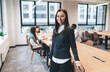 © BullRun - Young businesswoman in glasses standing beside desk, confidently smiling at camera, posing in casual-smart outfit in coworking space with team working in background