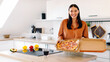 © Home-stock - Happy European lady holding open takeaway box with pizza, looking and smiling at camera, standing in kitchen interior, free space. Food delivery concept