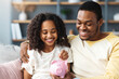 © Prostock-studio - Black girl and father putting coin into piggy bank, sitting on sofa at home. Happy african american dad teaching his cute daughter how to save money, living room interior. Financial education concept