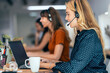 © BusinessClass by W61 - Smiling blond businesswoman talking through headset sitting by colleagues at desk in office