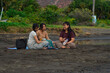 © agratitudesign - With the sun rising behind them, three teenage girls sit close on a beach mat, laughing and chatting comfortably in the soft morning light.