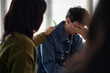 © Seventyfour - Teenage Caucasian boy sitting with hand covering face, showing distress during counseling session, female counselor placing hand on his shoulder, other teenagers present in background