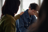 Teenage Caucasian boy sitting with hand covering face, showing distress during counseling session, female counselor placing hand on his shoulder, other teenagers present in background