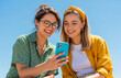 © Alingga - Two young women smile brightly while looking at a phone together under a clear and sunny blue sky.