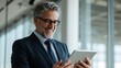 © DESIGN MOSAI - Experienced businessman in suit and glasses smiling while looking at a tablet device